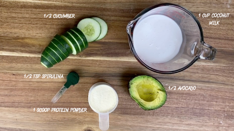 A flat lay of healthy smoothie ingredients: half a cucumber, one cup of coconut milk, half a teaspoon of spirulina, one scoop of protein powder, and half an avocado, neatly arranged on a wooden surface.