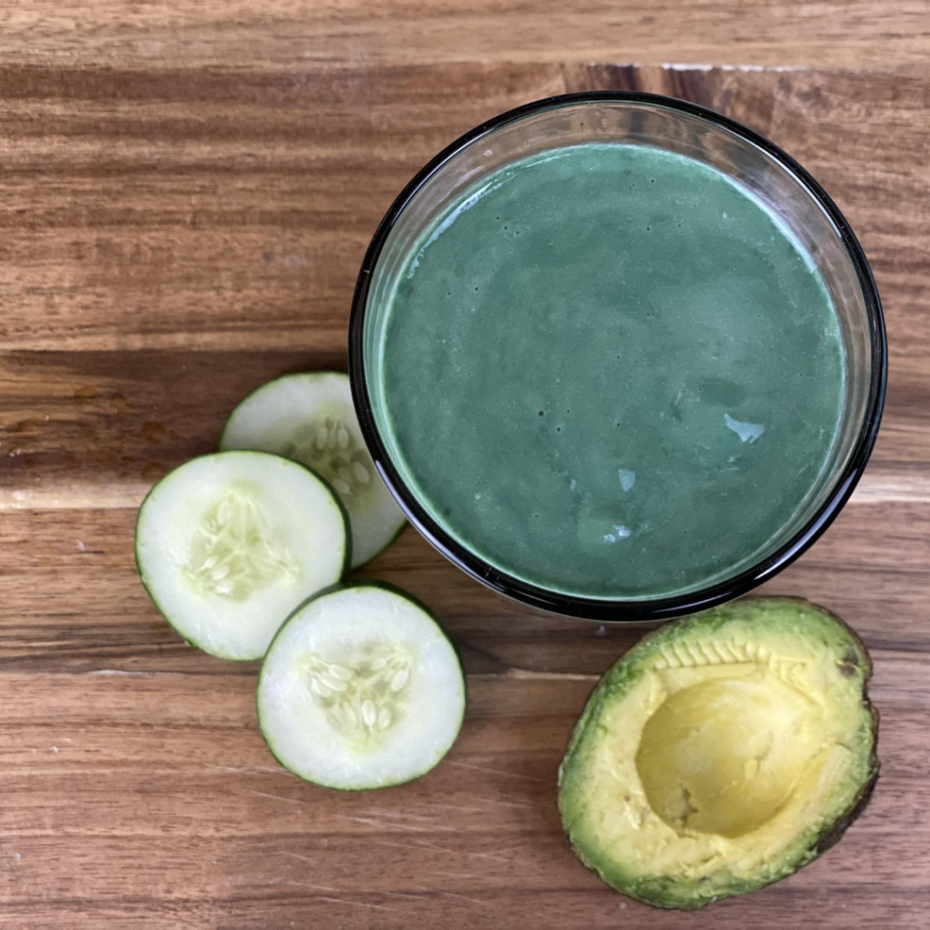 A fresh and healthy green smoothie accompanied by slices of cucumber and half an avocado on a wooden surface.