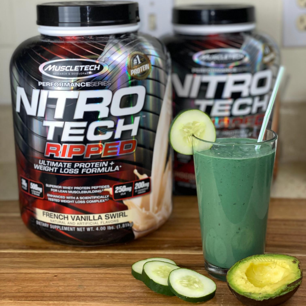 A nutritious post-workout setting with a glass of green smoothie, flanked by a sliced avocado and cucumber, with containers of muscletech nitrotech ripped protein supplement in the background.