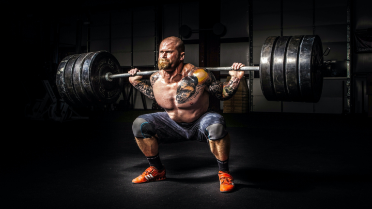 A tattooed athlete executing a heavy barbell squat with determination and focus in a gym setting.