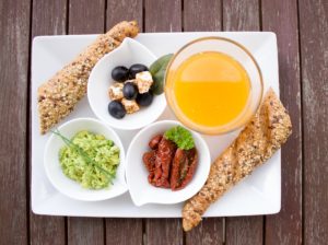 A nutritious breakfast platter with avocado spread, sun-dried tomatoes, olives with cheese, whole grain breadsticks, and a glass of orange juice served on a wooden table.