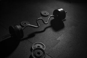 A black and white photo capturing an unoccupied curved barbell and loose weight plates on a dark gym floor, suggesting a pause in a workout session or the quiet atmosphere of a gym after hours.