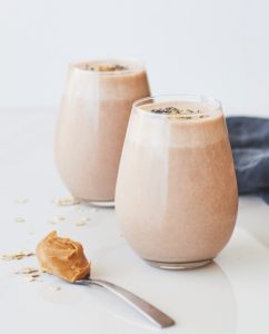 Two glasses of creamy smoothie topped with oats, with a dollop of peanut butter on a spoon in the foreground, suggesting a delicious and nutritious snack or breakfast option.