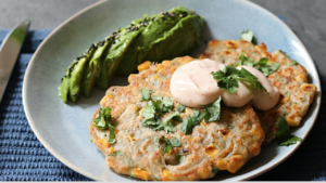 A plate featuring a savory corn pancake garnished with fresh herbs and a dollop of creamy sauce, accompanied by a slice of ripe avocado sprinkled with sesame seeds.