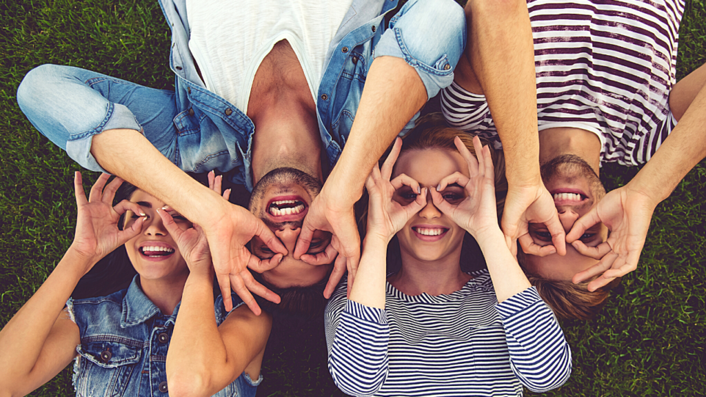 A group of cheerful friends lying on the grass, forming playful glasses shapes with their fingers while laughing and looking at the camera.