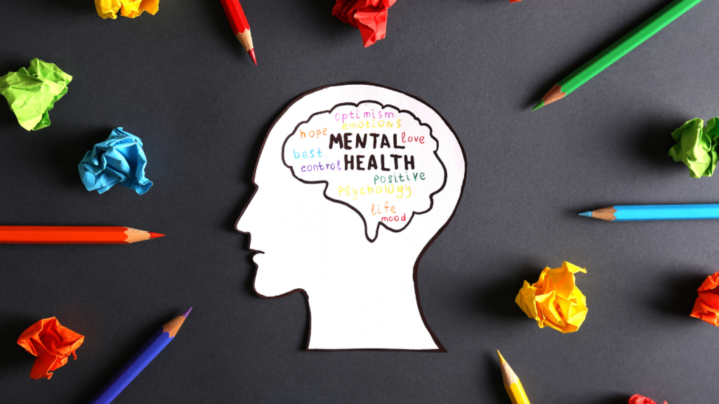 A creative paper cut-out silhouette of a head filled with positive mental health related words surrounded by colorful pencils and crumpled balls of paper on a dark background, symbolizing creativity and psychological well-being.