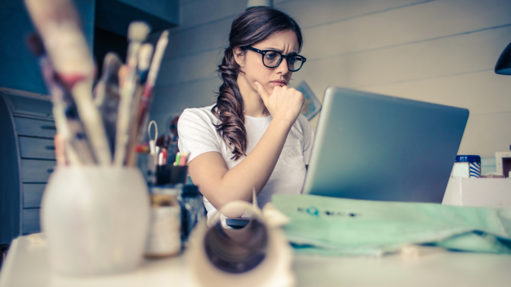 A thoughtful woman with glasses and a braid ponders deeply while looking at her laptop screen, surrounded by creative clutter and painting supplies.