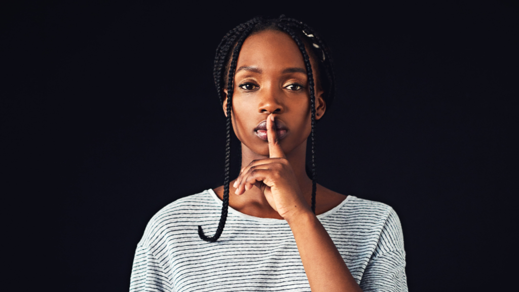 A person with braided hair placing a finger over their lips to gesture silence, set against a dark background.