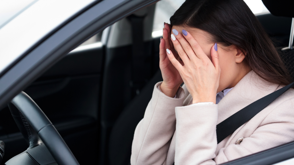 A stressed or upset woman sitting in the driver's seat of a car, covering her face with her hands.