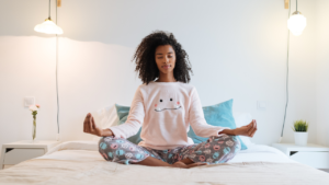 A serene woman practicing meditation on a bed in a well-lit, cozy bedroom setting.