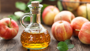 A clear glass bottle filled with golden apple cider vinegar, sealed with a cork, surrounded by fresh, ripe apples and green leaves on a rustic wooden table.