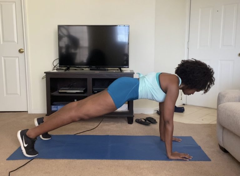 A woman engaged in a plank exercise on a blue mat in her living room, with a television set and various electronics in the background.