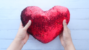 A pair of hands holding a large red sequined heart-shaped pillow against a white wooden background.