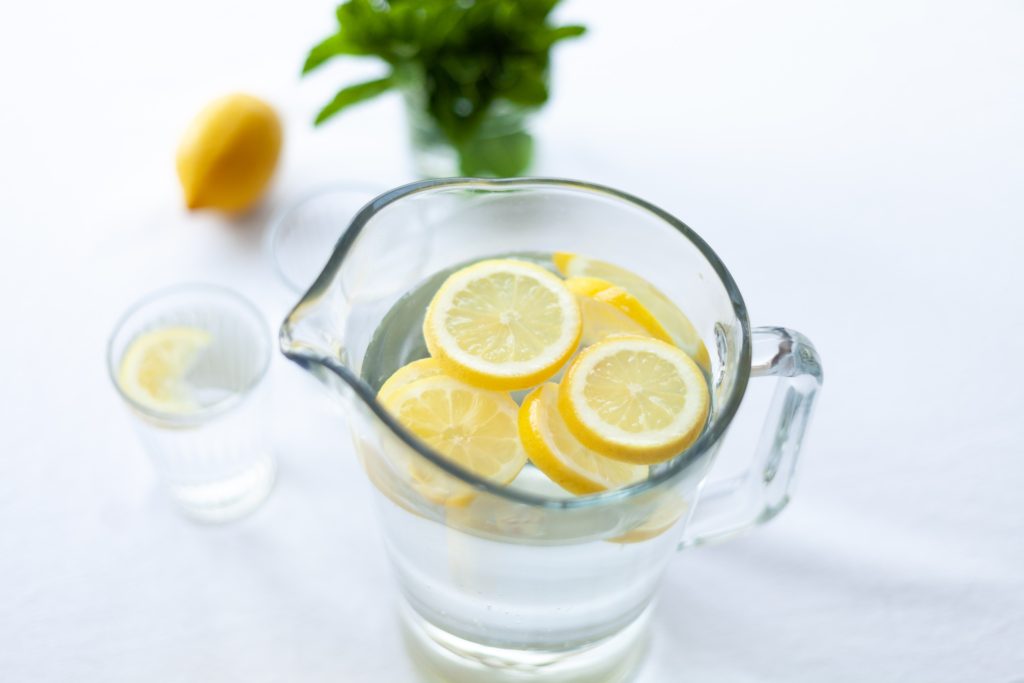 A refreshing pitcher filled with water and slices of lemon, accompanied by a glass and additional lemons in the background, suggesting a scene of summer refreshment or healthy hydration.