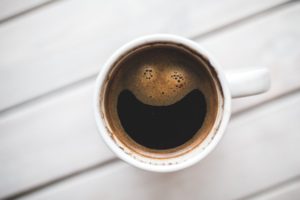 A top-down view of a white mug filled with coffee, showcasing a whimsical foam pattern that resembles a smiley face on the surface.