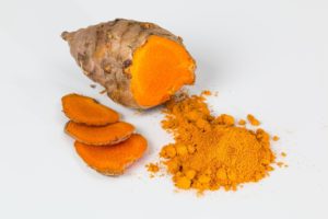 Fresh turmeric root partially sliced with bright orange, vibrant flesh on a white surface, alongside a small pile of turmeric powder.