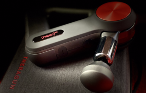 A close-up view of a theragun massage device, showcasing its sleek design and distinctive red and black color scheme, resting on a dark surface.