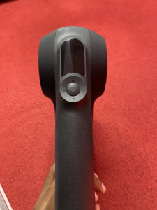 A close-up view of a handheld device with a trigger, possibly a barcode scanner or similar electronic tool, being held against a red carpeted background.