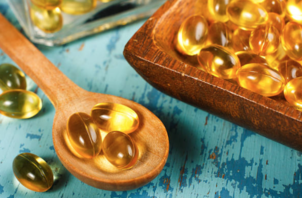 A wooden spoon rests on a distressed blue surface, holding several translucent golden capsules, while more capsules scatter around and a glass container lies in the background—suggesting a focus on dietary supplementation.