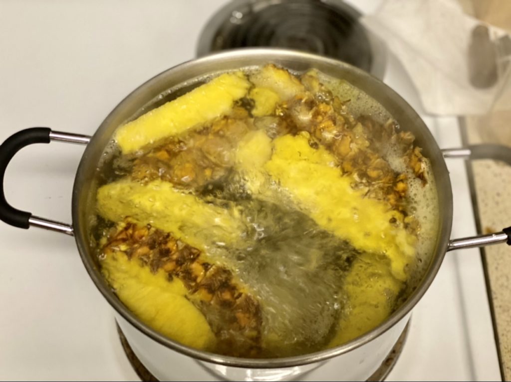 A pot of boiling water with corn on the cob and what appears to be pineapple boiling on the stove.