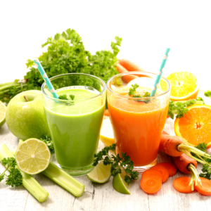 Two refreshing glasses of green and orange juice surrounded by a colorful assortment of fruits and vegetables, including apples, carrots, limes, and oranges, indicating a healthy and vibrant selection of fresh ingredients.