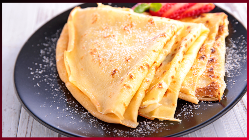 Golden crepes dusted with powdered sugar, accompanied by fresh strawberries on a black plate, ready for a sweet indulgence.