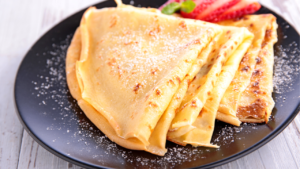 Golden crepes dusted with powdered sugar, accompanied by fresh strawberries on a black plate.