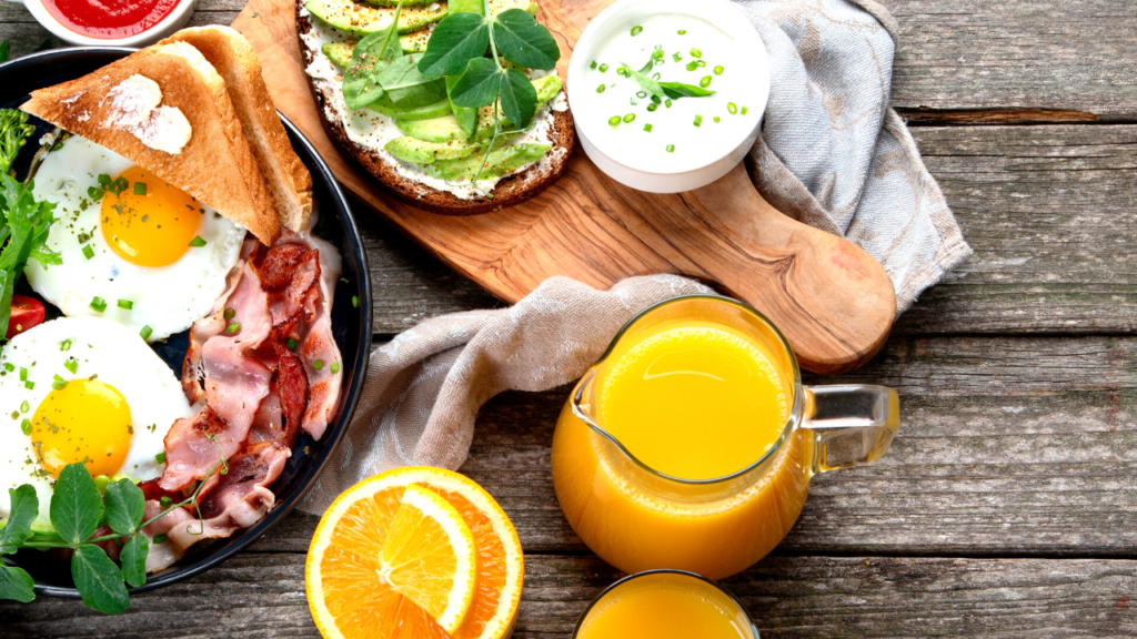 A hearty breakfast spread with sunny-side-up eggs, crispy bacon, avocado toast, fresh orange juice, and a sprinkle of herbs on a rustic wooden table.