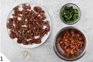 A variety of prepped ingredients laid out on a light surface, including a plate of sliced steak, a bowl of chopped green vegetables, and a bowl of diced tomatoes, suggesting the preparation phase for qu