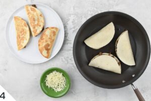 Golden-brown steak quesadillas on a white plate next to a skillet with more quesadillas cooking, accompanied by a small bowl of shredded cheese.