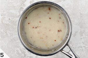 A top-down view of a saucepan containing a creamy queso sauce with specks of red seasoning on a speckled grey background.