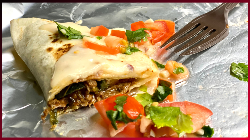A savory burrito cut in half to reveal its steak filling, topped with queso fondue and fresh diced tomato and herbs, ready to be enjoyed with a fork on standby.