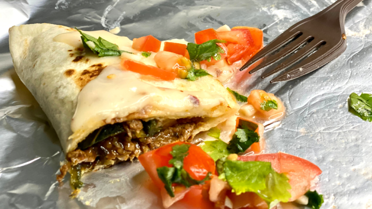 A half-eaten cheesy steak quesadilla garnished with fresh tomato and cilantro on a foil-lined plate, with a fork to the side, inviting a flavorful bite.