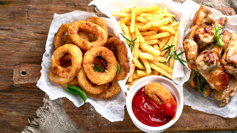 A tempting spread of fried onion rings, golden french fries, and grilled chicken drumsticks served with a side of ketchup, presented on a rustic wooden table.