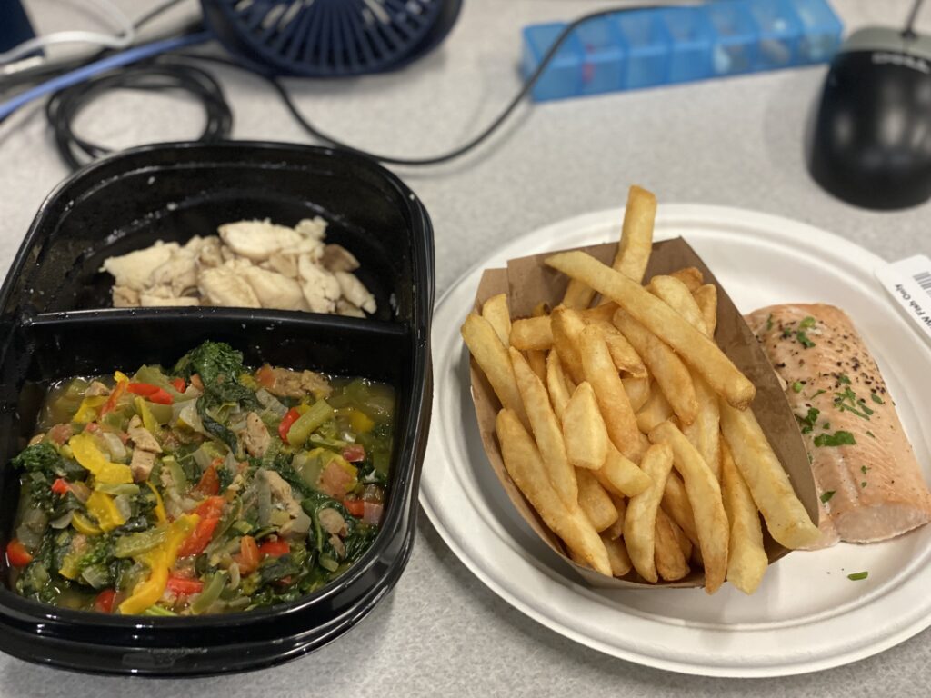 A balanced workspace lunch featuring a salmon filet with french fries on a paper plate, and a plastic meal prep container filled with a broccoli and beef stir fry.