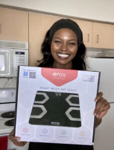 A smiling woman showing off her new smart body fat scale in a kitchen setting.