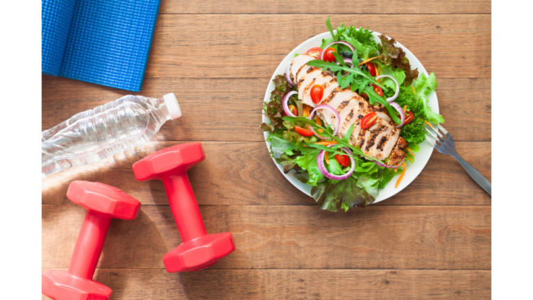 A healthy lifestyle concept with a fresh salad, grilled chicken breast, a pair of red dumbbells, a water bottle, and a yoga mat on a wooden surface.