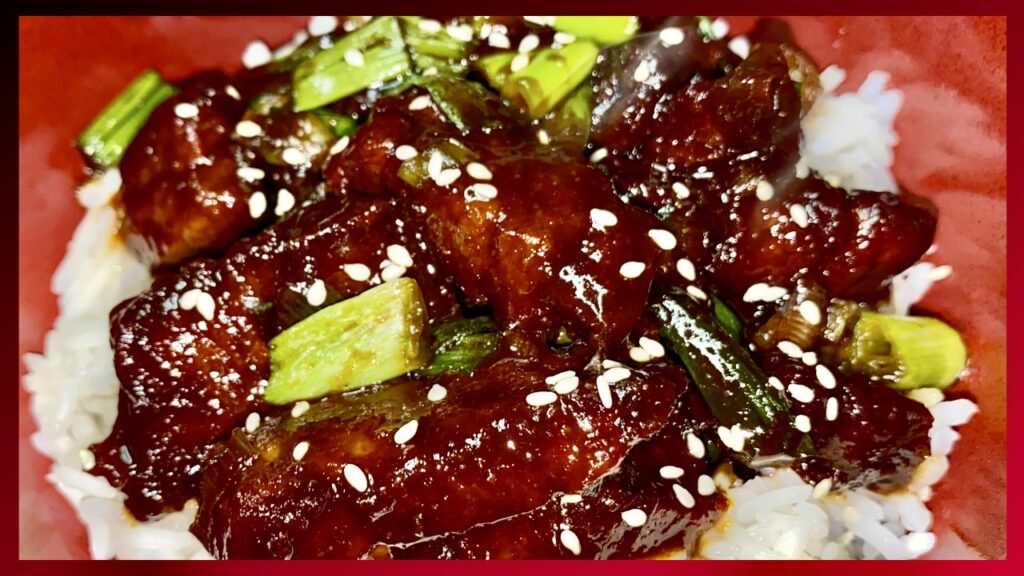 A close-up view of a Mongolian Beef recipe featuring glossy, sauce-coated chunks of meat garnished with green onions and sesame seeds, served over a bed of white rice in a red bowl.