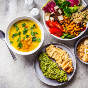 A colorful and nutritious vegetarian meal spread featuring a bowl of chickpea soup garnished with herbs, alongside dishes of quinoa salad, roasted vegetables, avocado toast, and hummus on bread from the 15 Plant-Based Recipes - eBook.