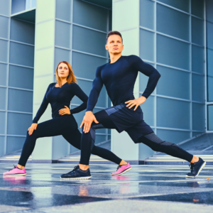 Two people in athletic wear doing a stretching exercise in an urban setting.