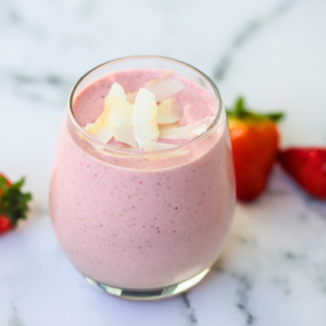 A creamy strawberry smoothie topped with coconut flakes in a clear glass, with fresh strawberries in the background.