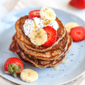A stack of fluffy pancakes topped with fresh slices of banana and strawberry, a dollop of cream, and a sprinkle of chia seeds, served on a blue plate with more berries on the side from the Vegan Recipes - eBook.