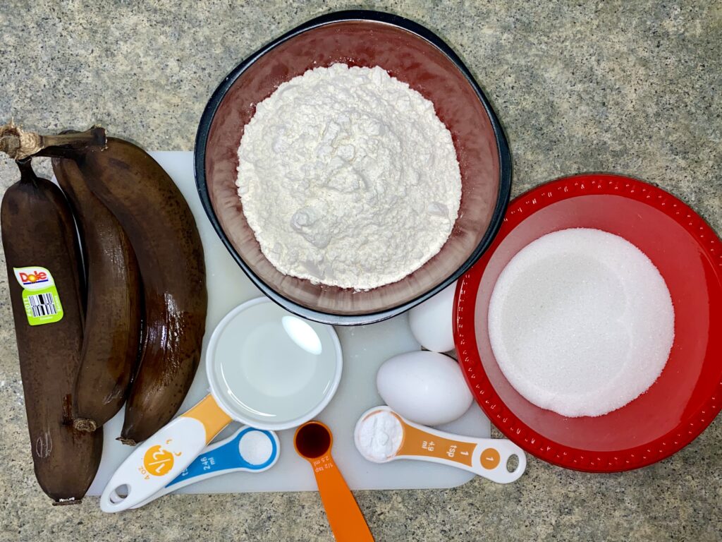 Ingredients for baking on a kitchen counter, including flour, sugar, bananas, eggs, and measuring spoons.