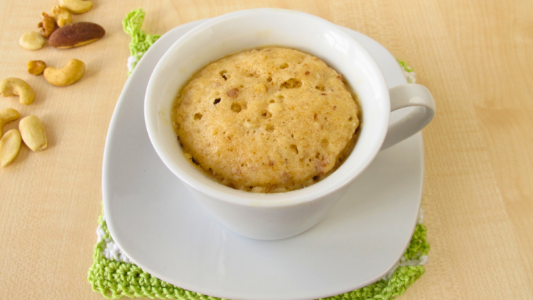 A freshly-baked, fluffy mug cake served in a white cup on a green coaster, surrounded by scattered cashew nuts and an almond on a light wooden surface.