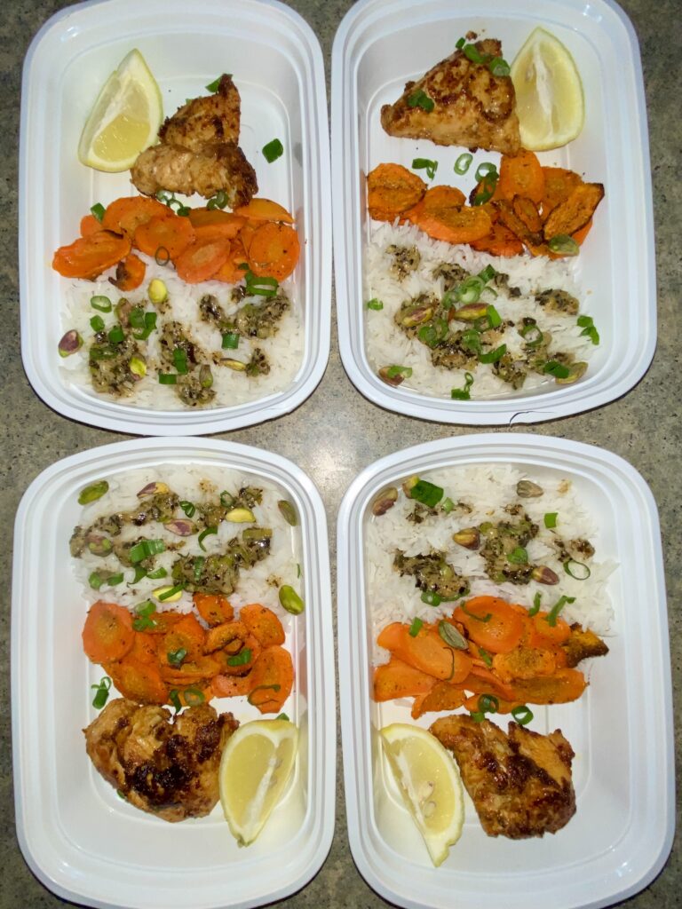 Four meal prep containers with a balanced meal of grilled chicken, rice with lentils, steamed carrots, and a lemon wedge for a zesty flavor boost.