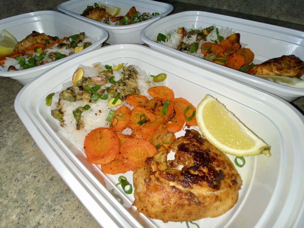 Prepared meal containers showcasing a nutritious dinner with grilled chicken, seasoned rice, sliced carrots, and a wedge of lemon.