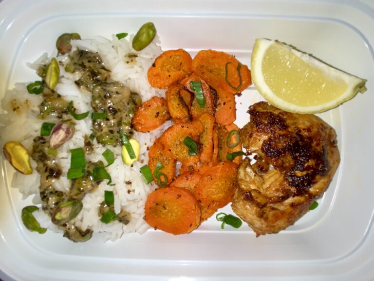 A well-balanced meal consisting of grilled chicken with a lemon wedge, rice garnished with herbs and nuts, and sliced sautéed carrots, neatly arranged in a white compartmentalized meal container.