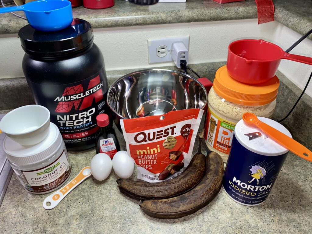 Assorted ingredients for a protein-packed snack: supplement powders, a quest bar, eggs, banana, and coconut oil gathered on a counter, ready for a nutritious recipe.