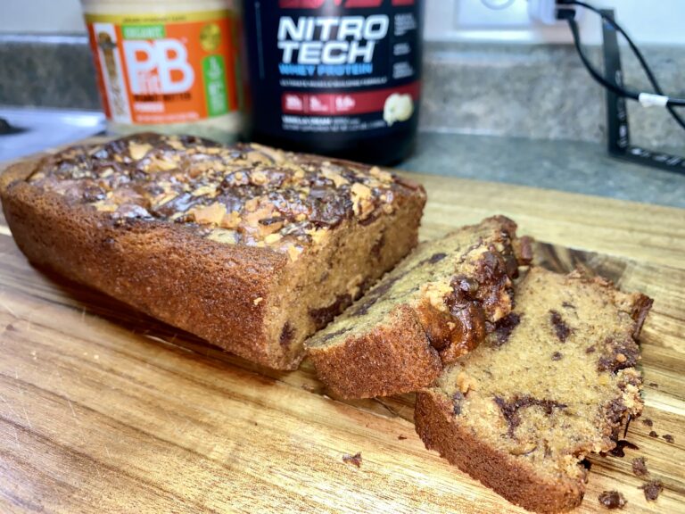 A freshly baked loaf of banana bread with nuts and chocolate chips, sliced and ready to enjoy, sitting on a wooden cutting board with a jar of peanut butter and a container of whey protein in the background, suggesting a blend of indulgence and health-consciousness in the kitchen.