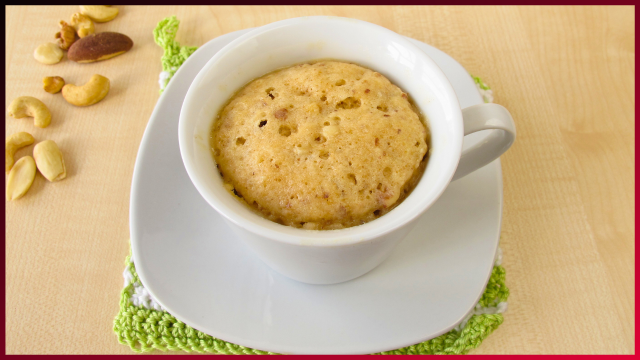 A freshly baked mug cake served in a white cup on a saucer, placed on a green knitted cloth with scattered nuts around on a wooden surface.